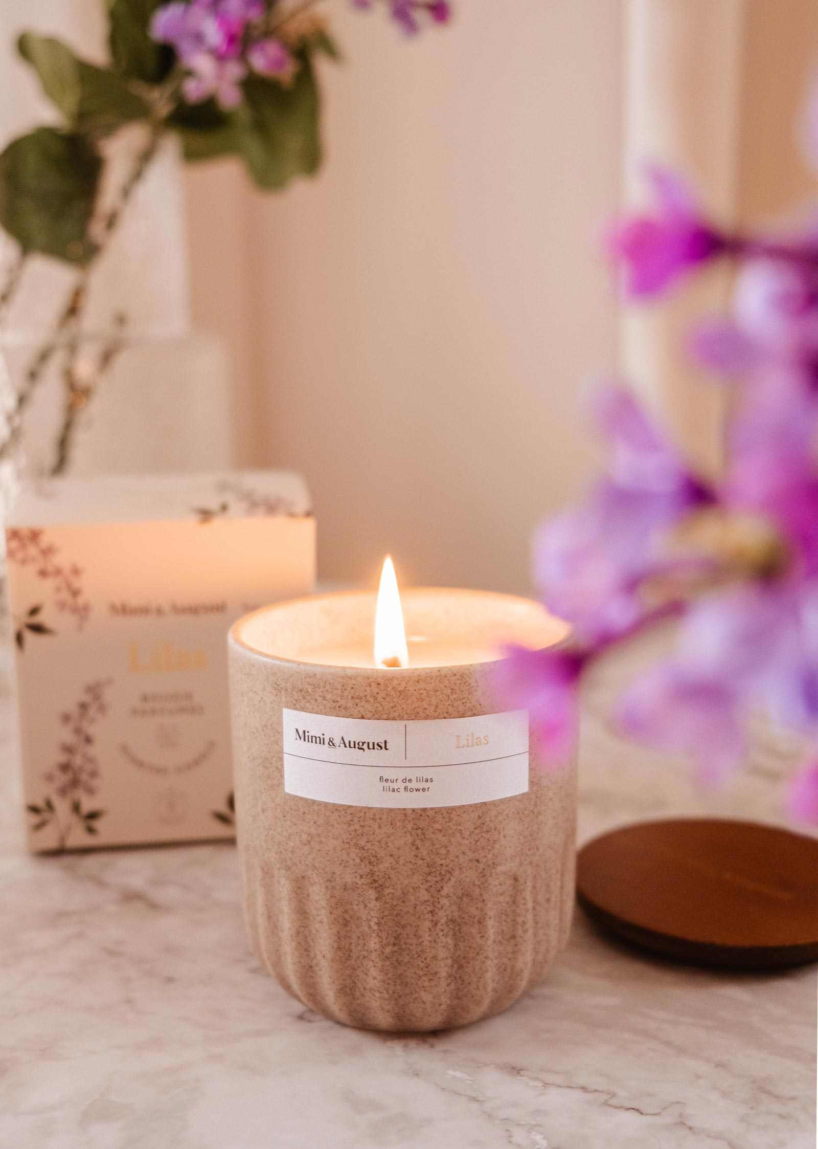 Candle with a label on a marble surface with flowers and packaging in the background mimi-and-august