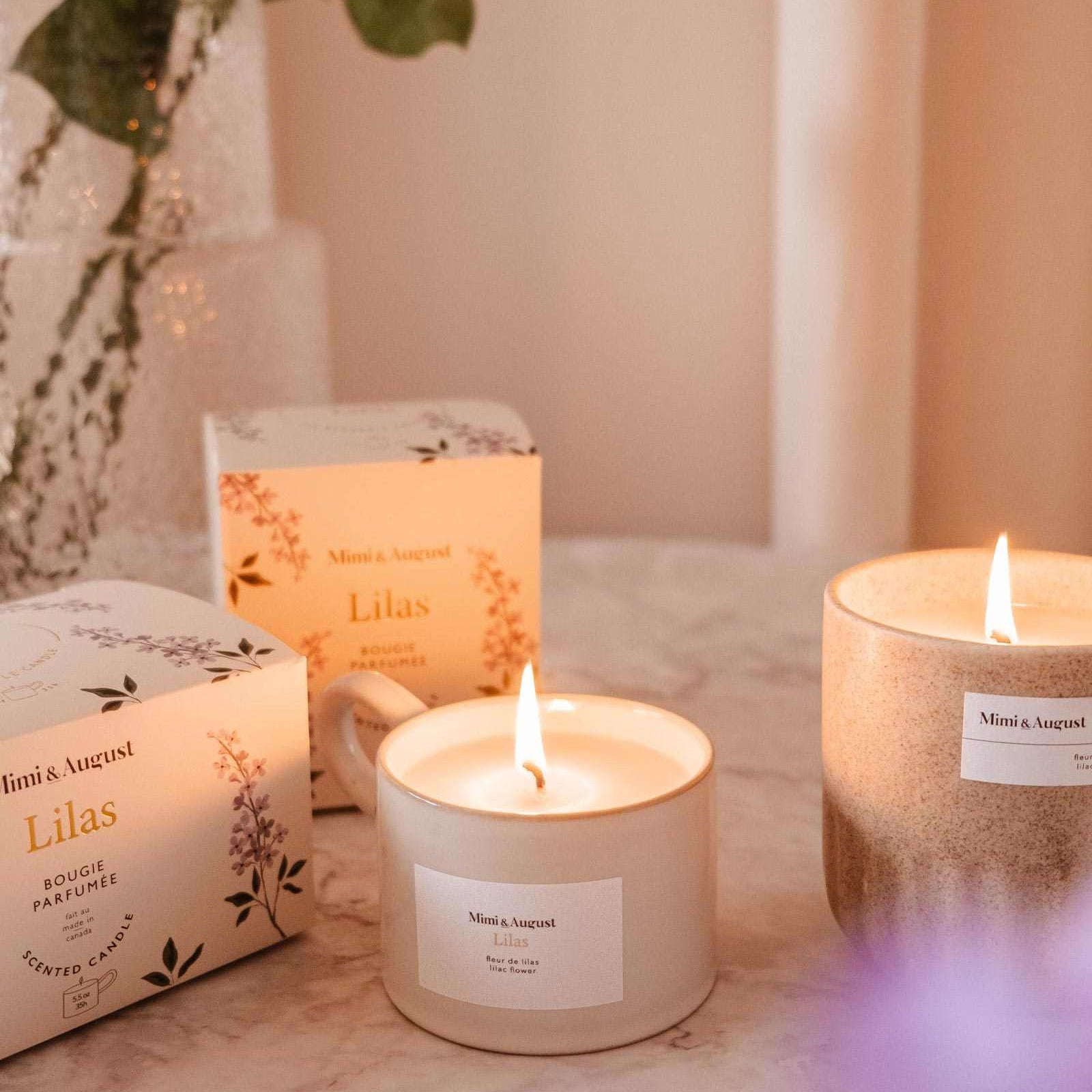 Candles and boxes with 'Ninon & August' branding on a surface with flowers in the background. mimi-and-august