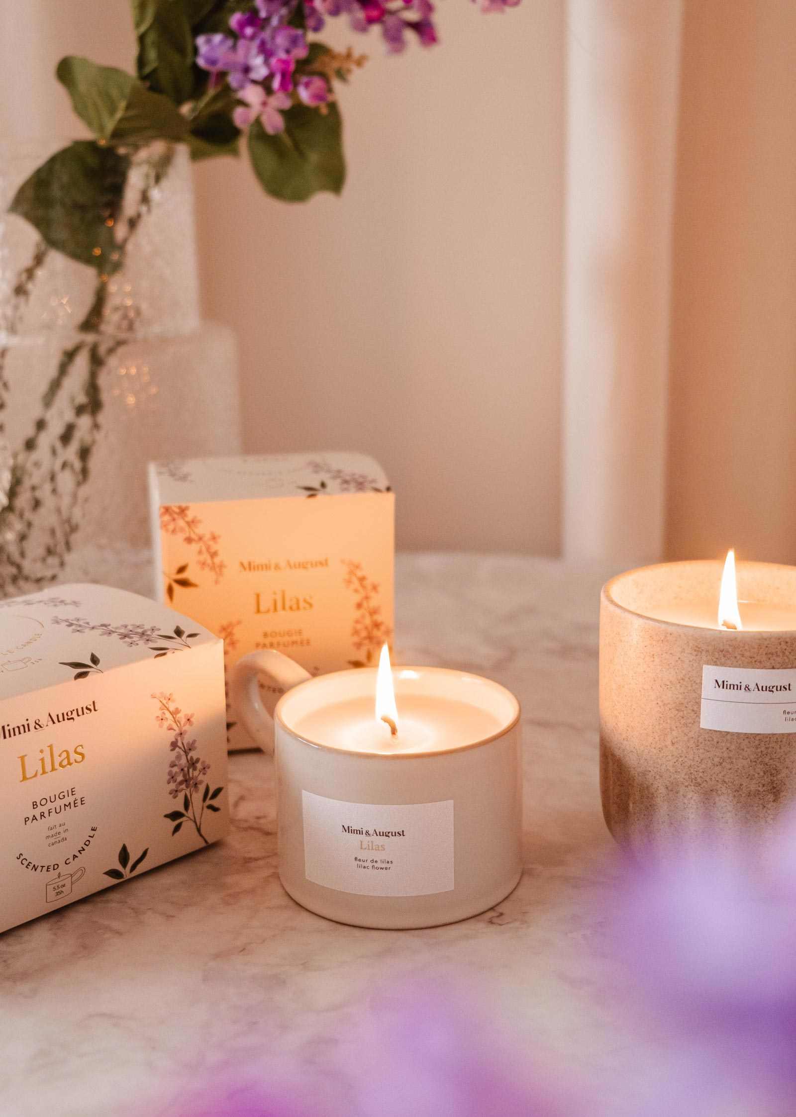 Candles and boxes with 'Ninon & August' branding on a surface with flowers in the background. mimi-and-august