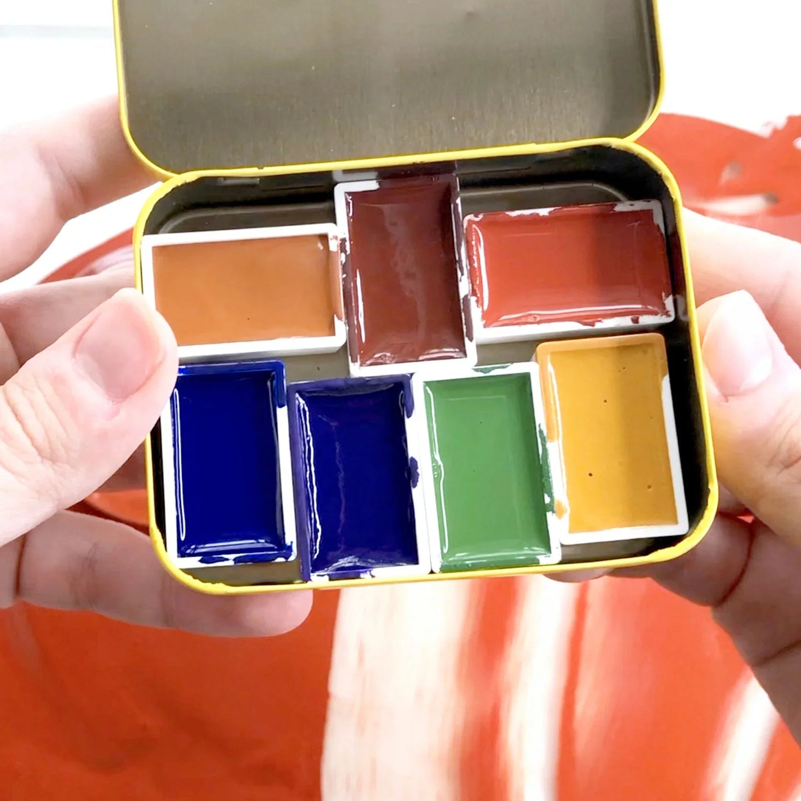 Hand holding a small open tin of watercolor paints with visible colors.