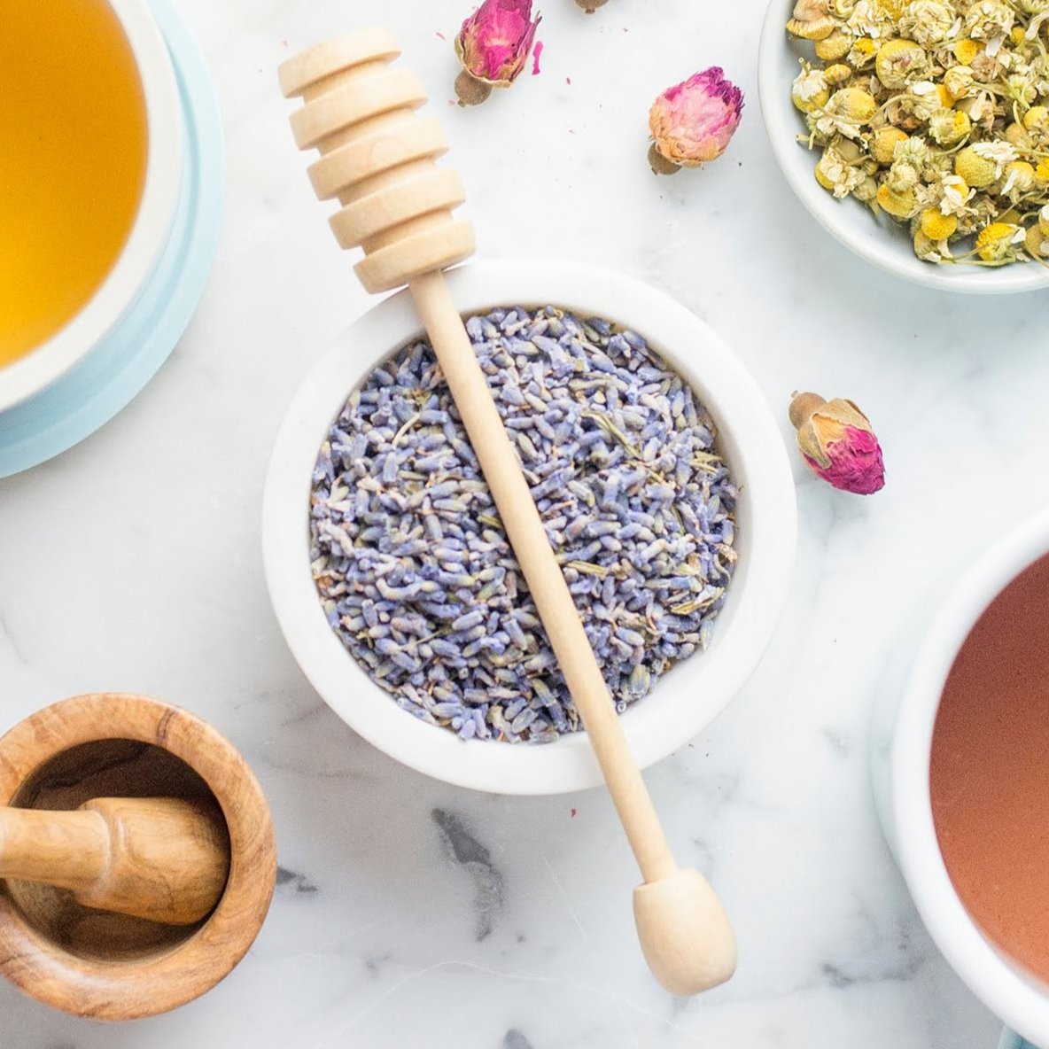 Bowl of lavender flowers with a wooden spoon on a marble surface, surrounded by tea cups and bowls.