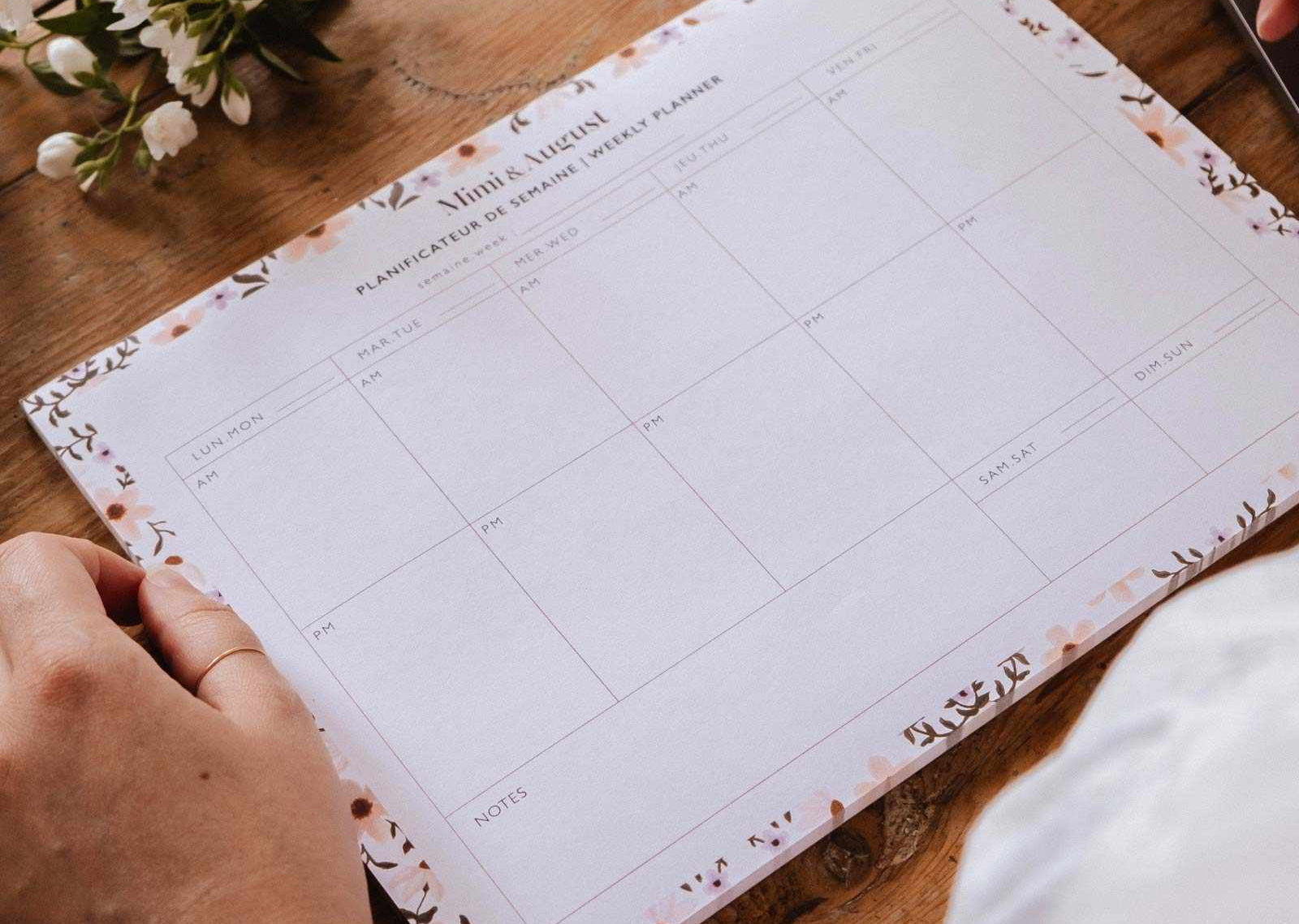 Person holding a planner on a wooden table with a laptop and flowers in the background