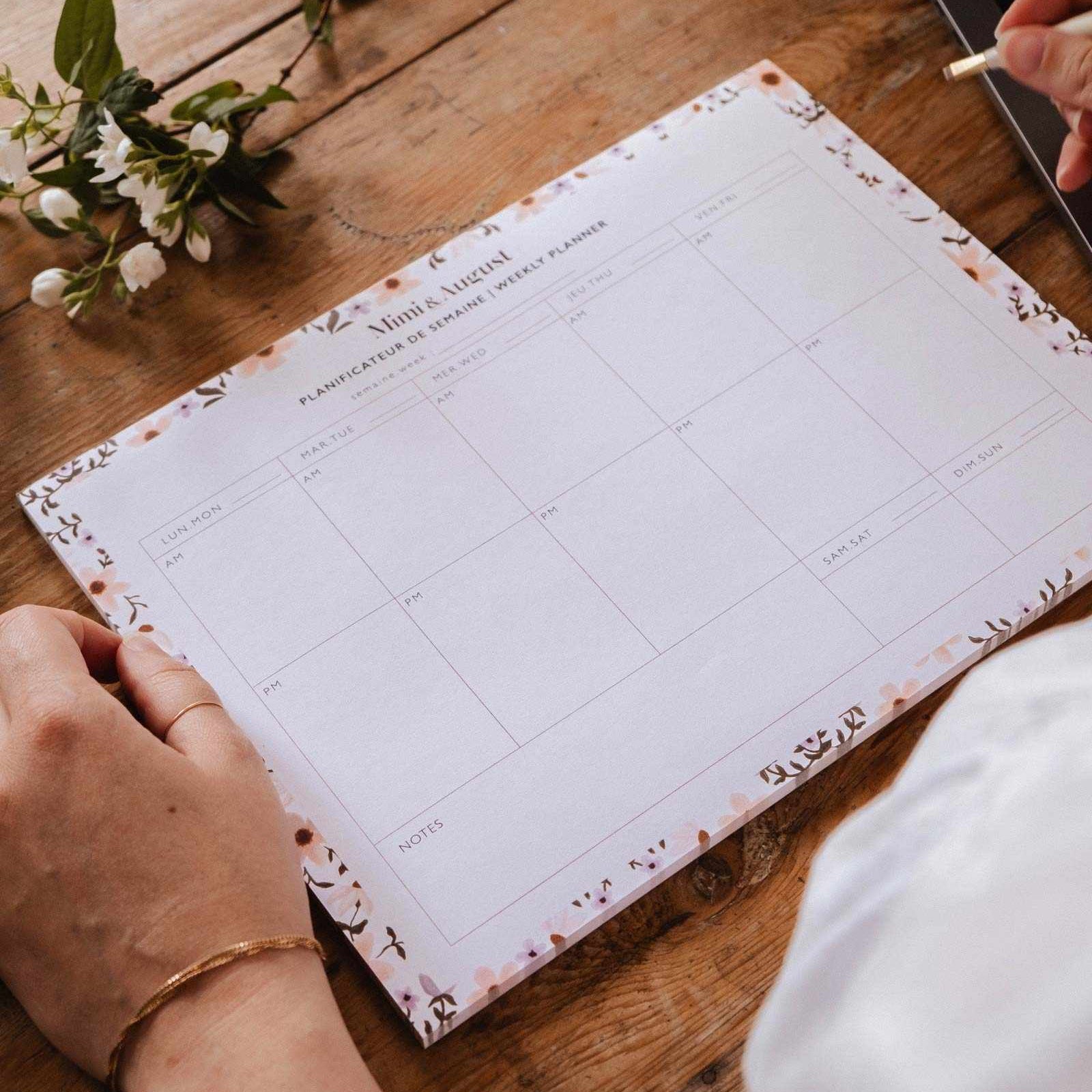 Person holding a planner on a wooden table with a laptop and flowers in the background