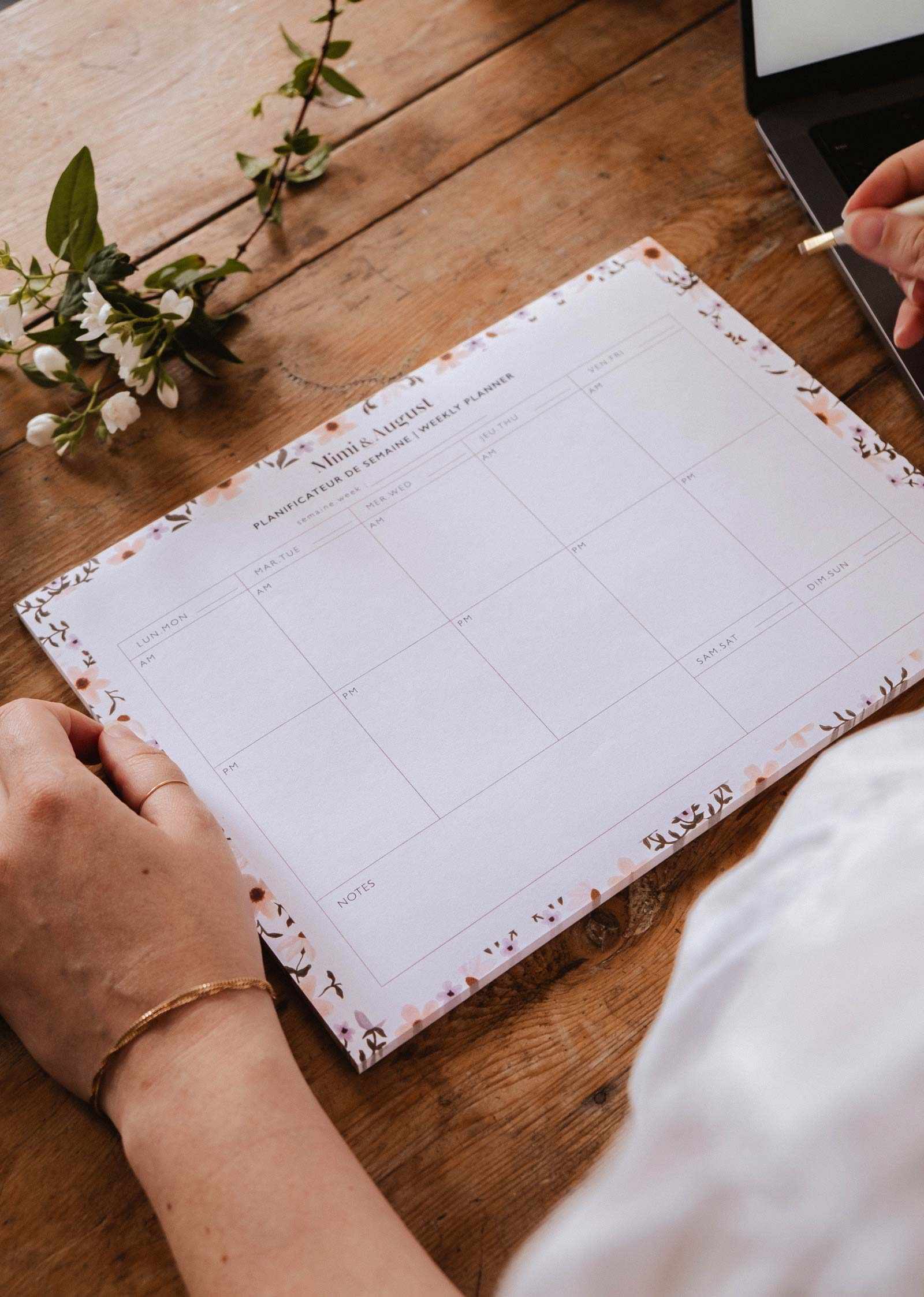 Person holding a planner on a wooden table with a laptop and flowers in the background