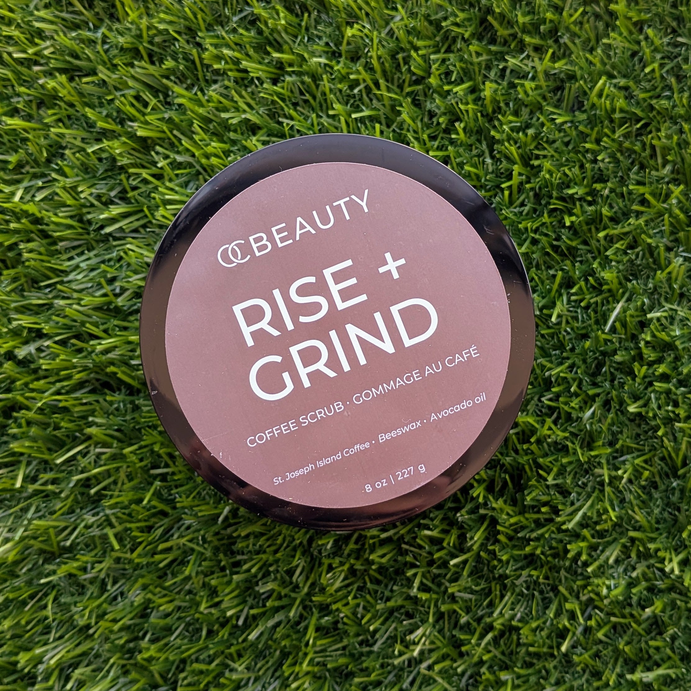 OC Beauty Rise + Grind Coffee Scrub, featuring a dusty rose-brown label with white text, placed on a green grass background.
