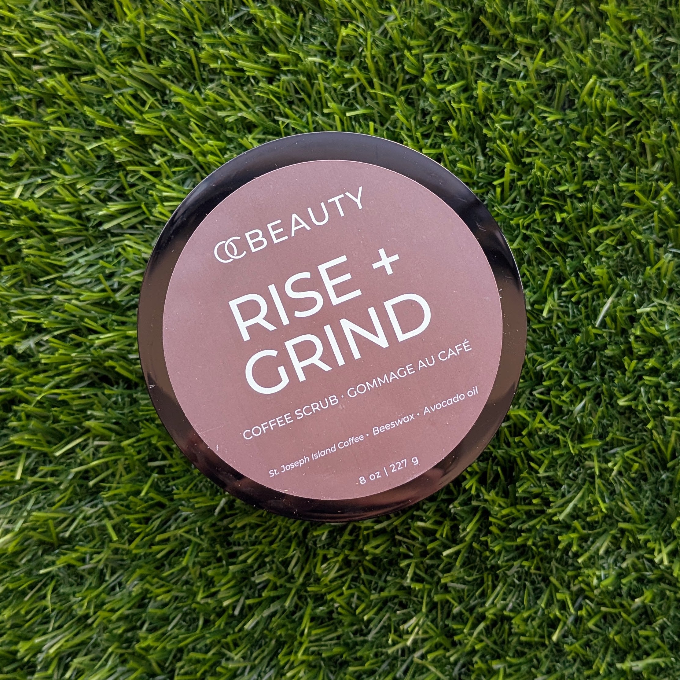 OC Beauty Rise + Grind Coffee Scrub, featuring a dusty rose-brown label with white text, placed on a green grass background.