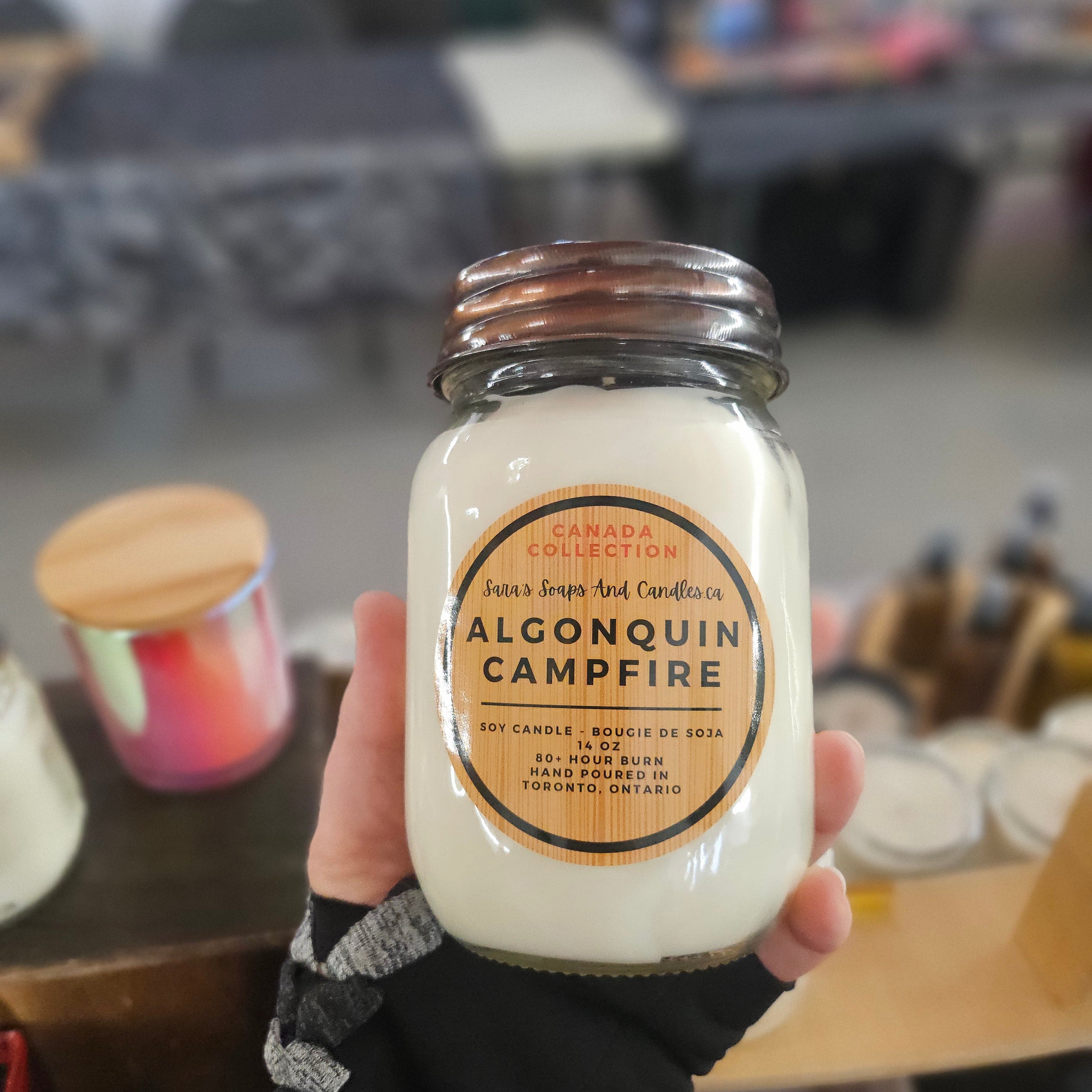 Hand holding a jar of Algonquin Campfire candle with a blurred indoor background from Stop and Smell the Candles