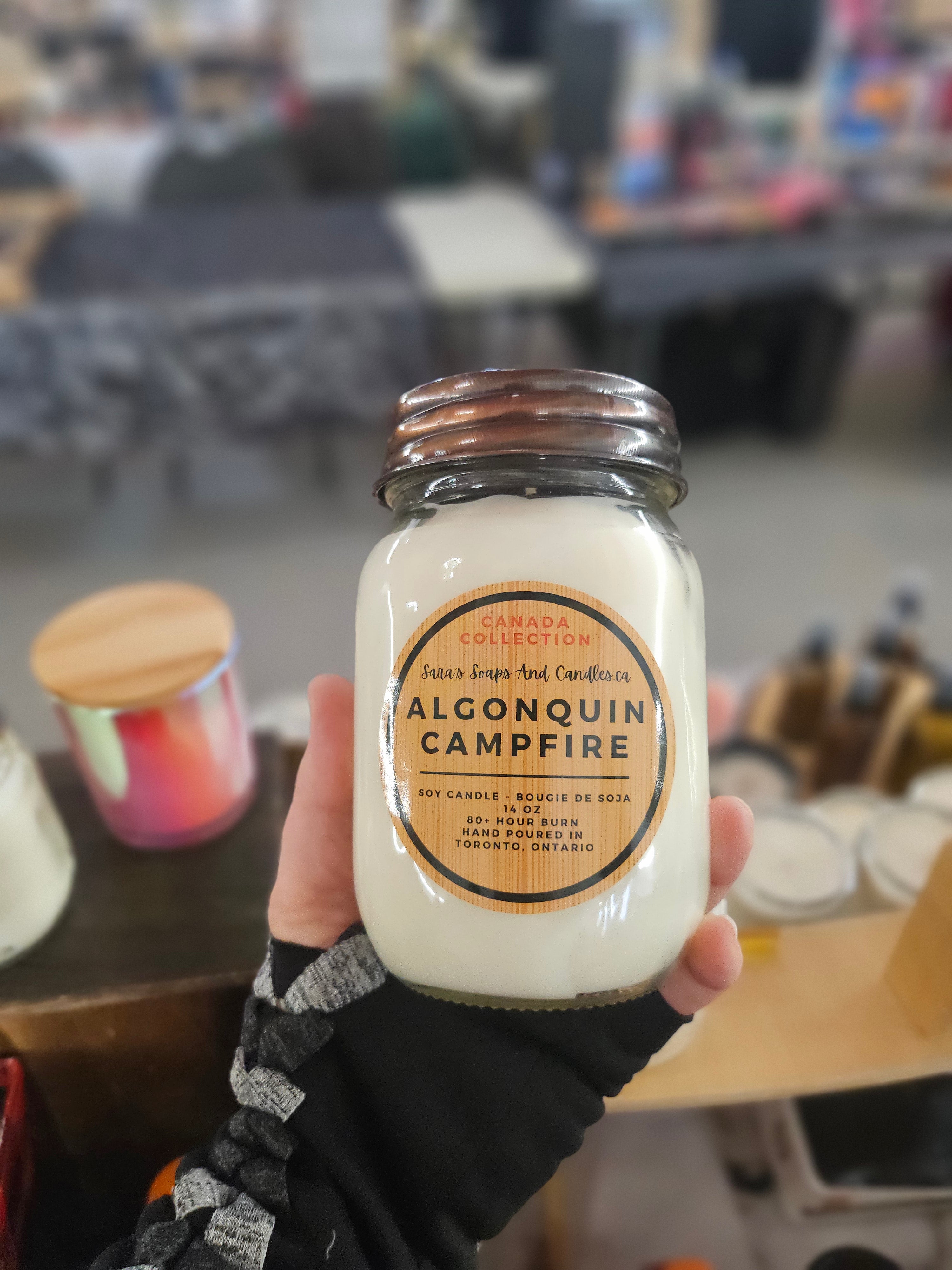 Hand holding a jar of Algonquin Campfire candle with a blurred indoor background from Stop and Smell the Candles