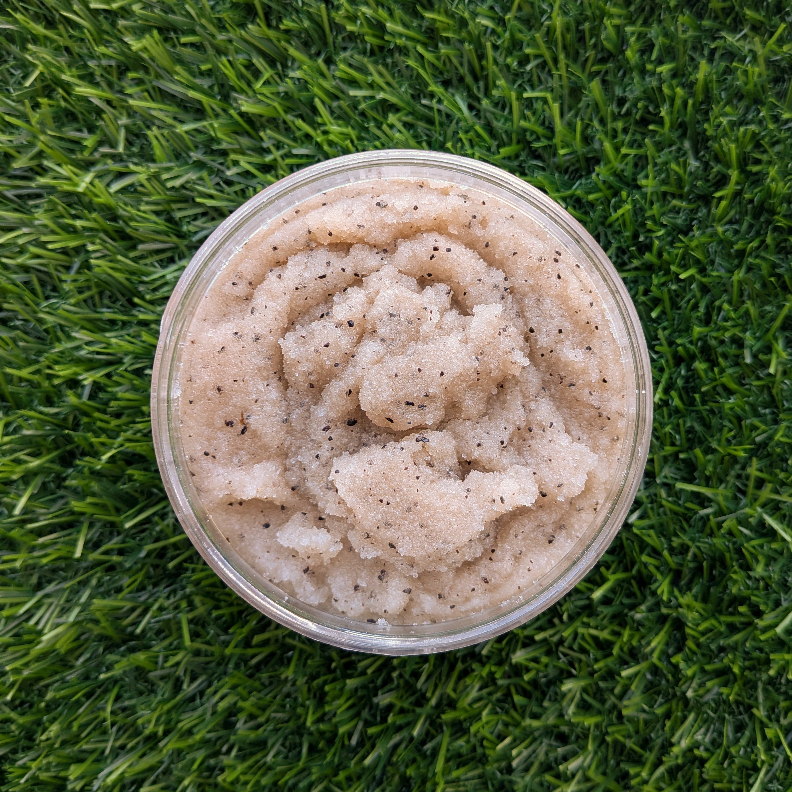 OC Beauty Rise + Grind Coffee Scrub  plastic tub filled with a light beige textured scrub containing small black specks, photographed from above on a green grass background.