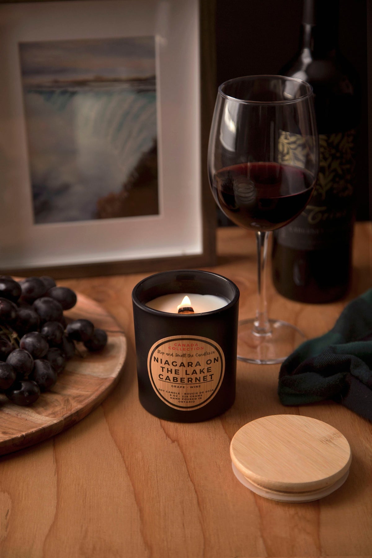 Stopandsmellthecandles Niagara on the Lake Cabernet soy candle – 8 oz black jar with lit wooden wick and wooden lid beside grapes and red wine