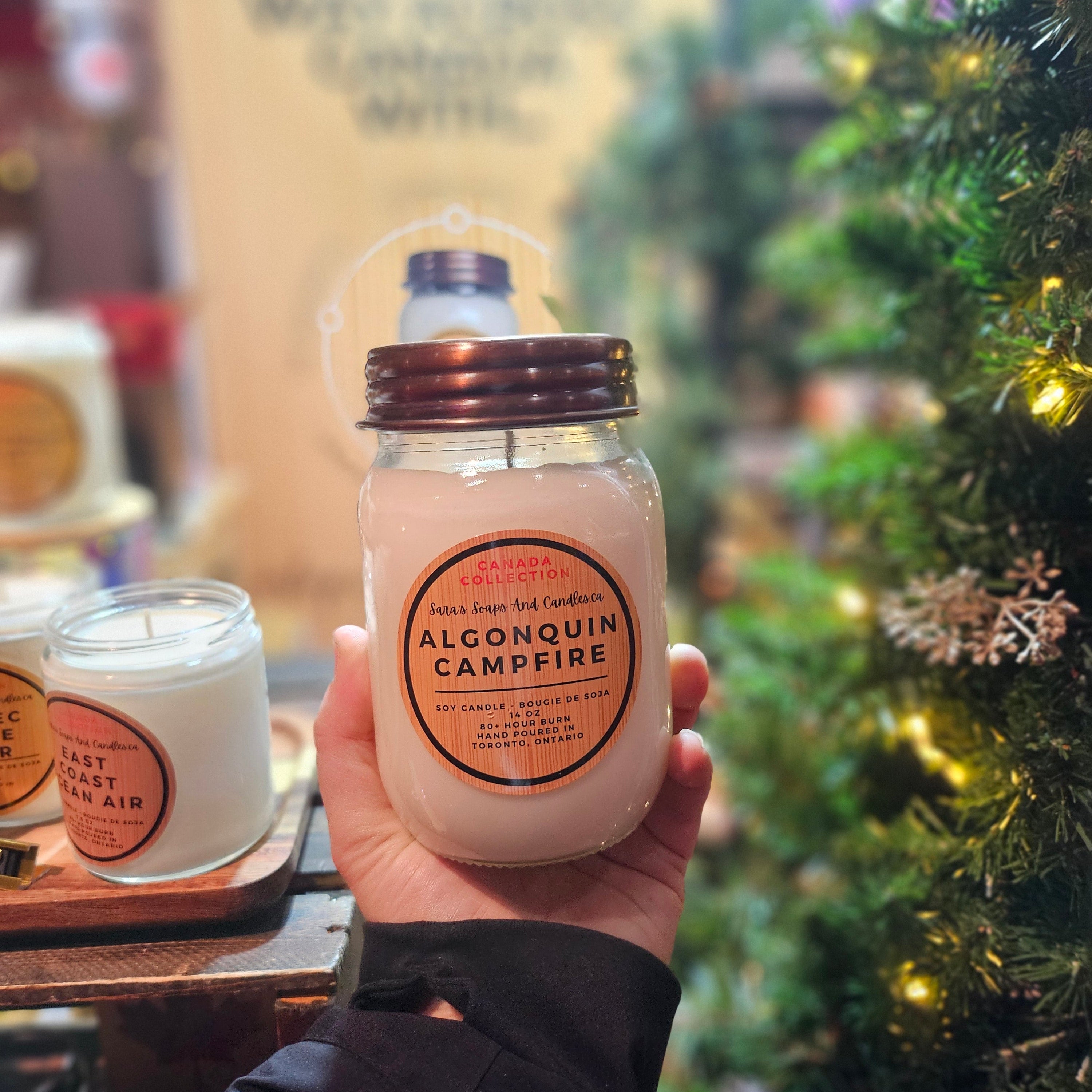 Hand holding a Algonquin Campfire candle in front of a Christmas tree from Stop and Smell the Candles