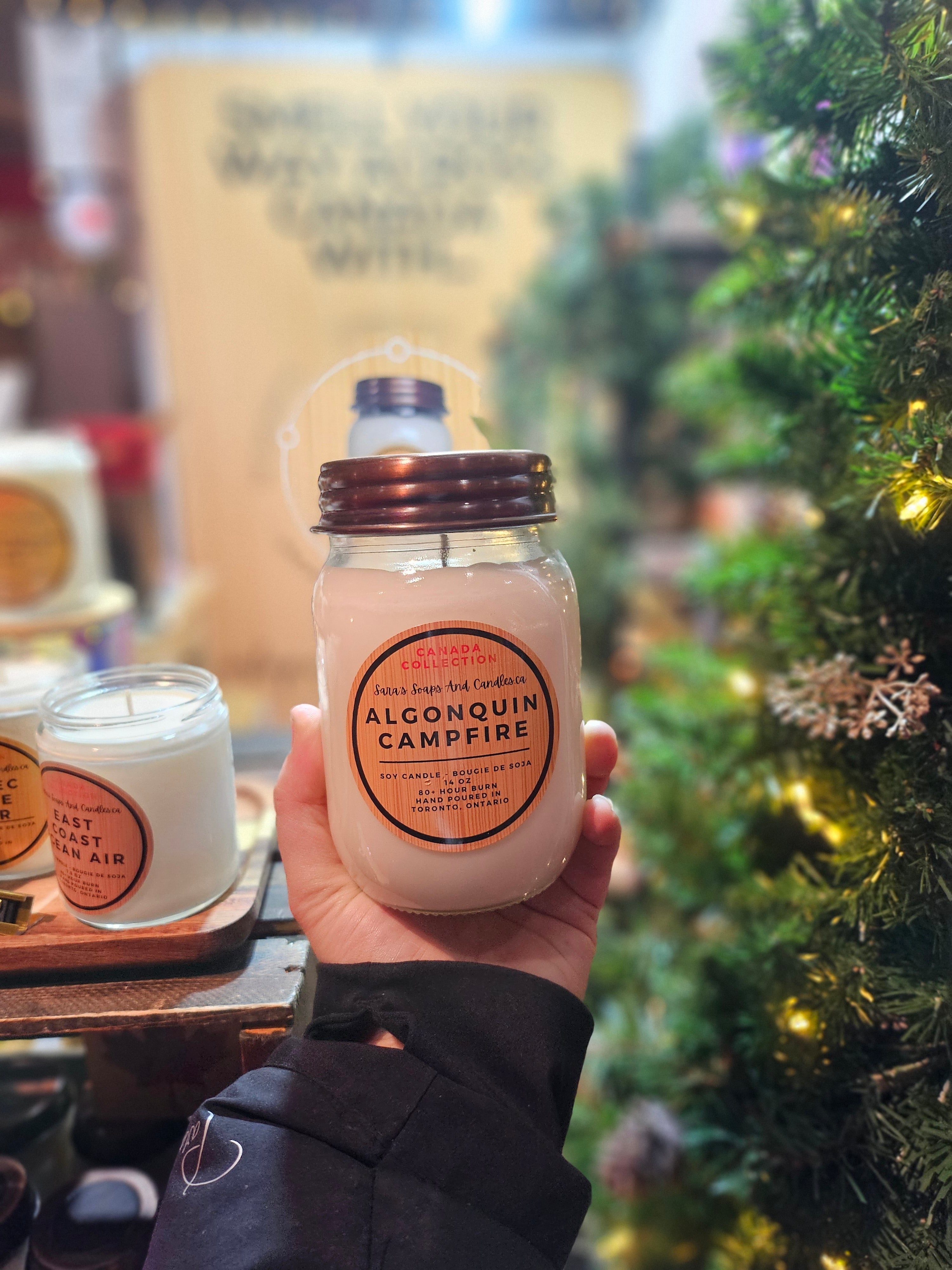 Hand holding a Algonquin Campfire candle in front of a Christmas tree from Stop and Smell the Candles