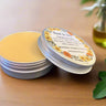 Balm in a tin with a decorative label on a wooden surface, surrounded by green leaves and a glass container.