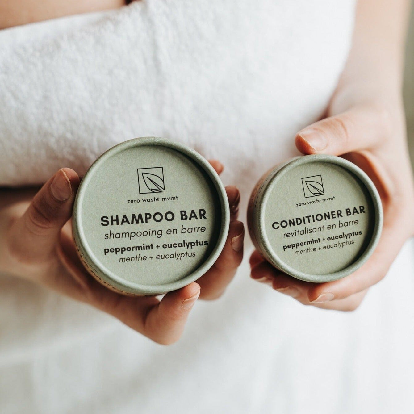 Two hands holding containers labeled 'Shampoo Bar' and 'Conditioner Bar' with a towel in the background.