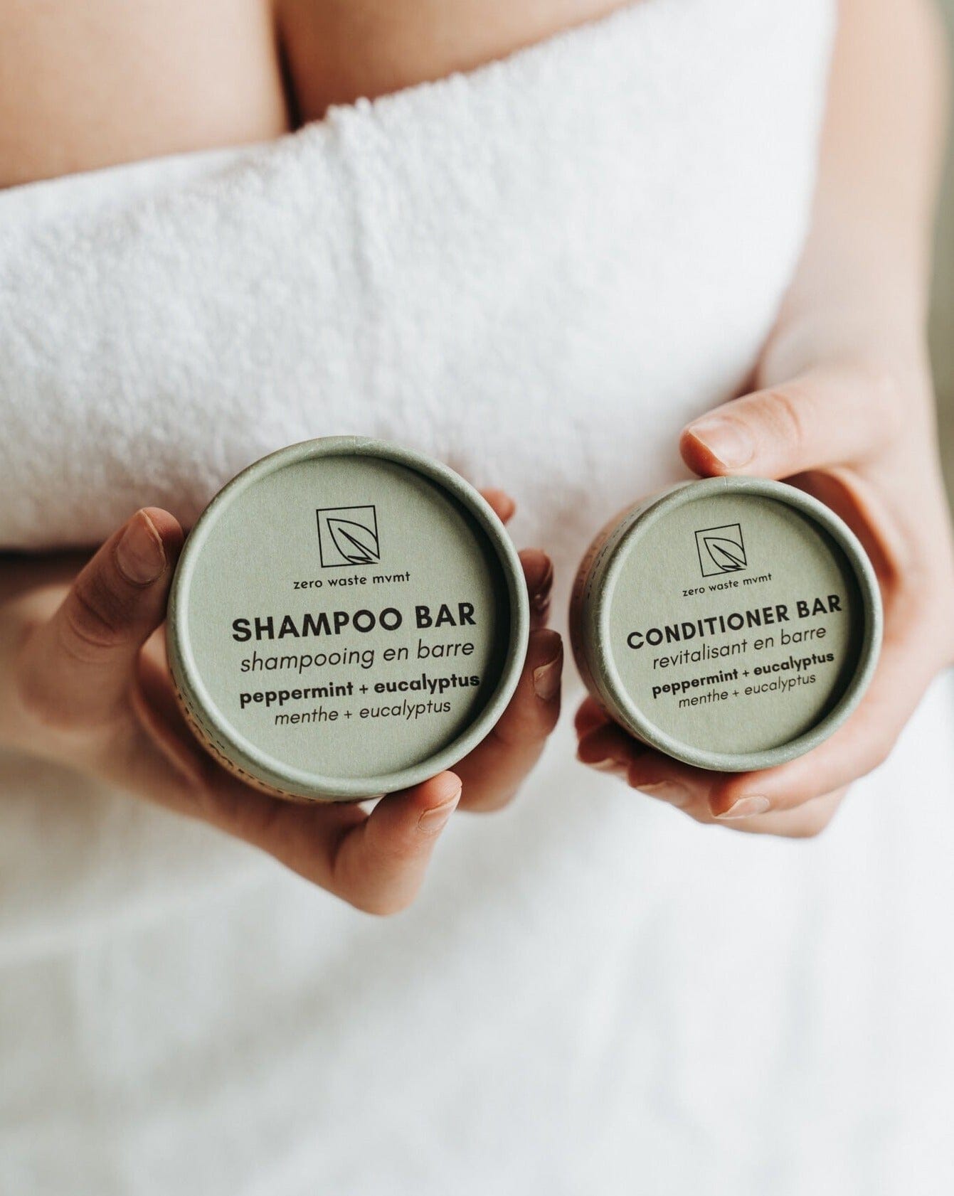 Two hands holding containers labeled 'Shampoo Bar' and 'Conditioner Bar' with a towel in the background.