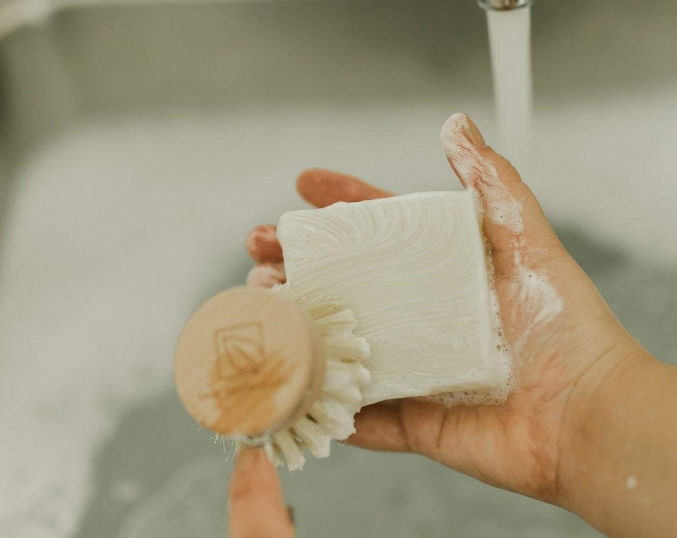 Hand holding a scrub brush and bar of soap under running water in a sink.