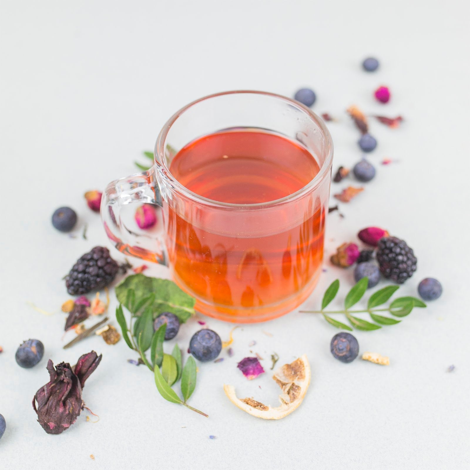 Clear glass mug filled with red tea surrounded by berries and herbs on a light background