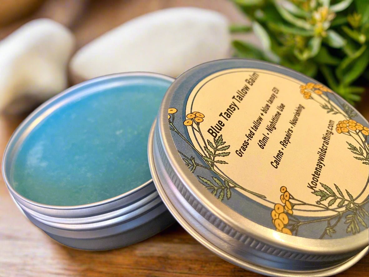 Open tin of Blue Tales Tallow Balm with label and blue balm inside, on a wooden surface with greenery.