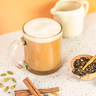 A frothy light brown beverage in a clear glass mug with a handle, set on a speckled white surface with cinnamon sticks, green cardamom pods, and a small wooden bowl of loose tea. A cream-colored pitcher is blurred in the background against a warm orange backdrop.
