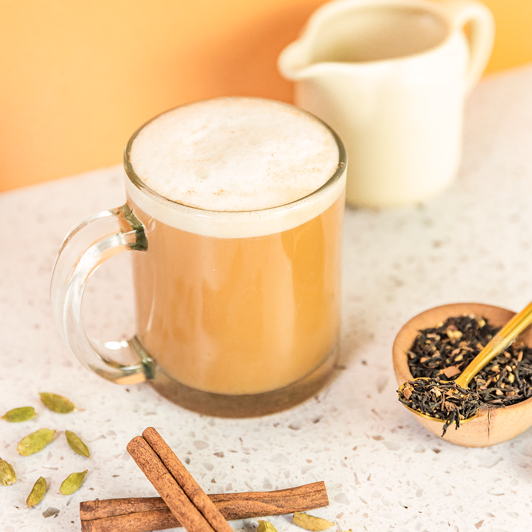 A frothy light brown beverage in a clear glass mug with a handle, set on a speckled white surface with cinnamon sticks, green cardamom pods, and a small wooden bowl of loose tea. A cream-colored pitcher is blurred in the background against a warm orange backdrop.