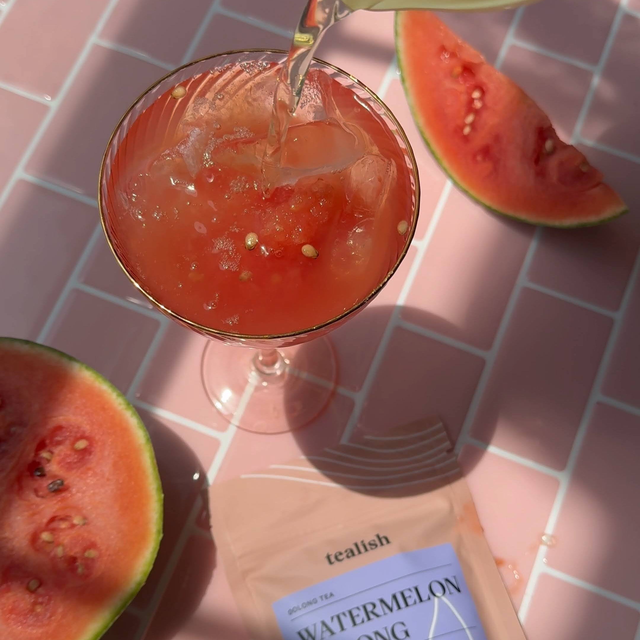 Cocktail being poured into a glass with watermelon slices and a tea bag labeled 'Watermelon Oolong' on a pink grid background.