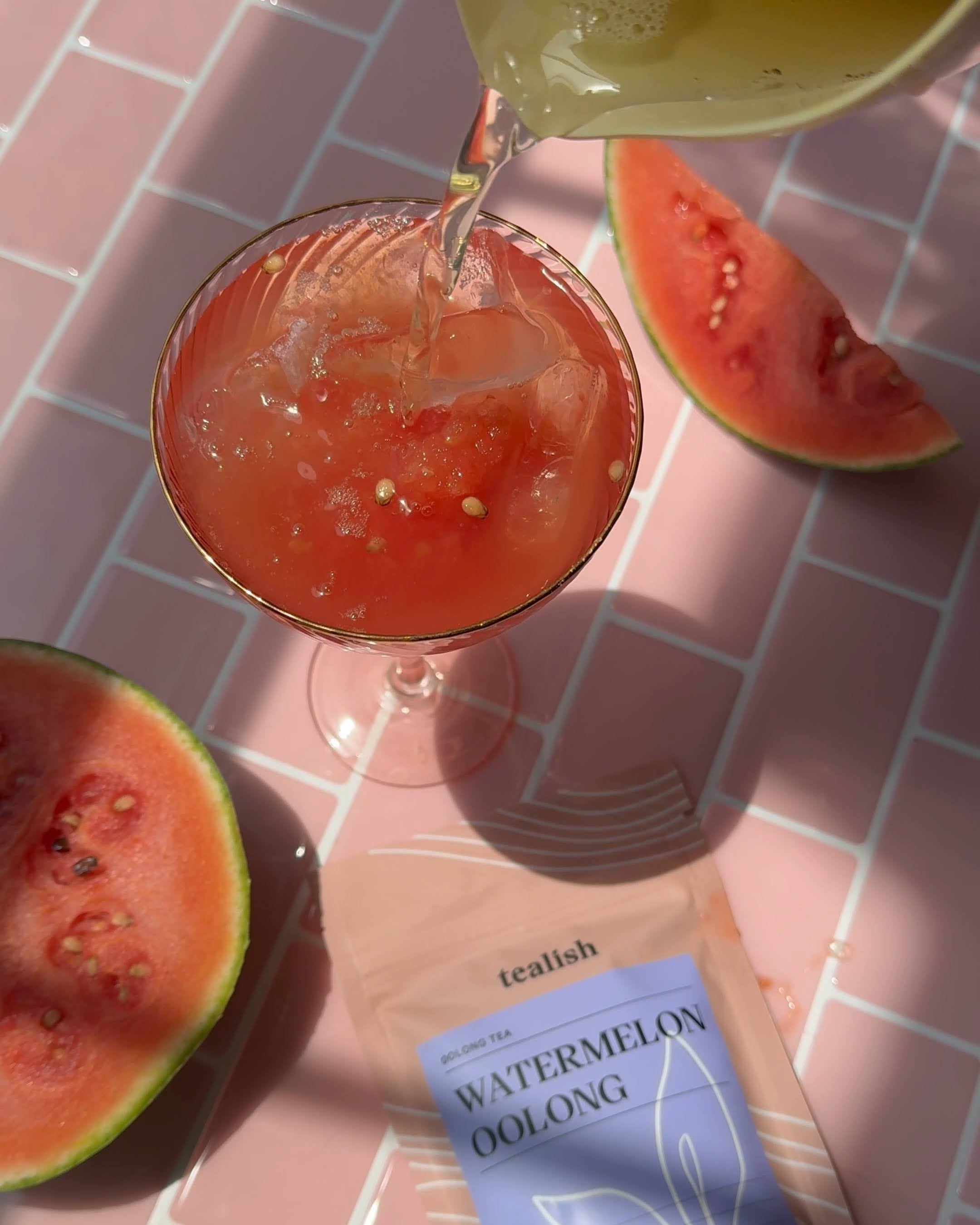 Cocktail being poured into a glass with watermelon slices and a tea bag labeled 'Watermelon Oolong' on a pink grid background.