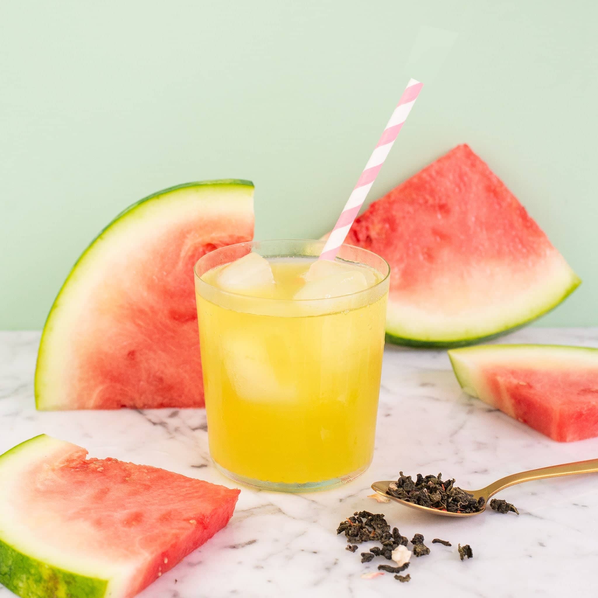 Glass of yellow liquid with a pink straw, surrounded by watermelon slices on a light green background